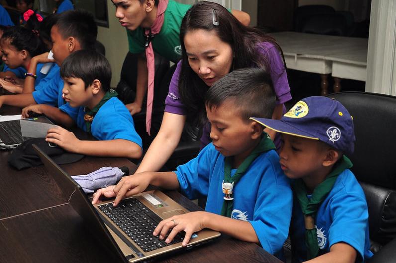 A group of scouts working on laptops during an indoor digital learning session. In the foreground, two young scouts in blue uniforms and neckerchiefs focus on a laptop screen, while an adult leader leans in to support them. More children and another leader are visible in the background.