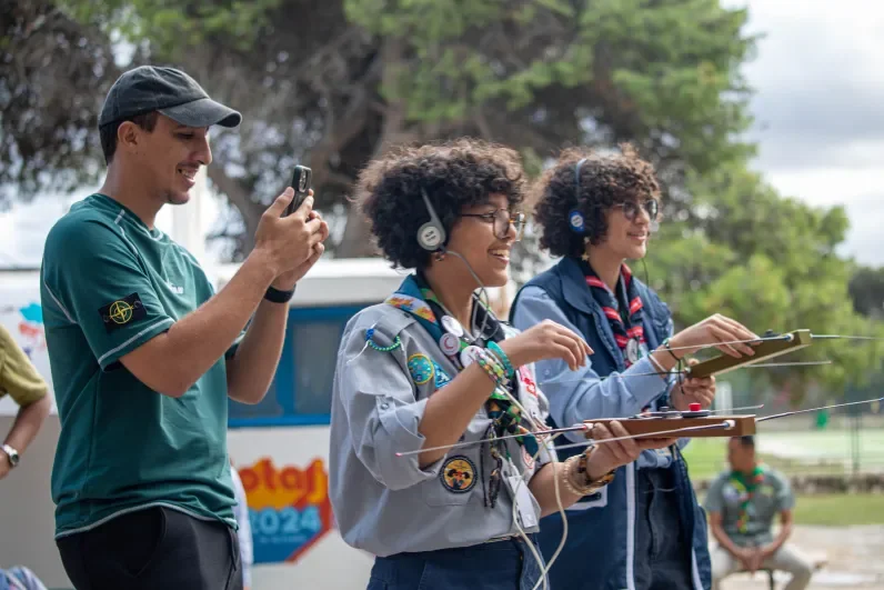 Three young scouts participating in an outdoor radio activity. Two of them are wearing headphones and holding direction-finding antennas, while a third is smiling and taking a photo with a smartphone. They appear engaged and happy. A JOTA-JOTI 2024 banner and trees are visible in the background.